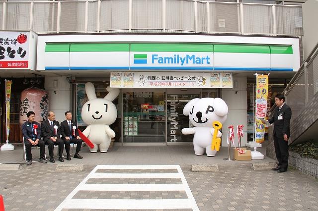 コンビニ前、左に三人の男性が座っており、出入口を挟んでマイナちゃんとマイキー君が立ち、右で男性が司会進行をしているオープニングセレモニーの様子の写真