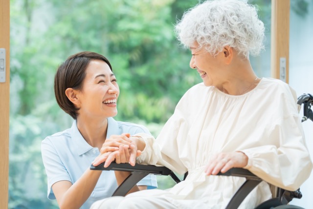 介護職員初任者研修イメージ画像 車いすに乗った高齢女性に、介護職員の女性が笑顔で寄り添う様子