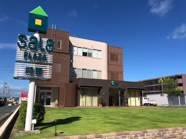 サーラプラザ湖西の外観写真 茶色を基調とした建物で、建物の前には緑の家をモチーフにした看板ときれいな芝生が広がっている。