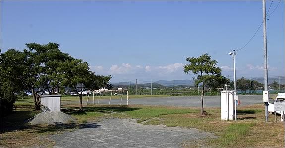 青い空の下、大きな樹木も立ち、遠くには山が見える自然豊かな運動広場の写真